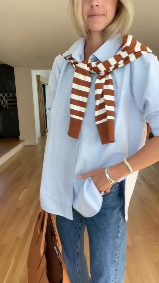 Woman in oversized light blue shirt, blue jeans, and striped sweater tied around shoulders.