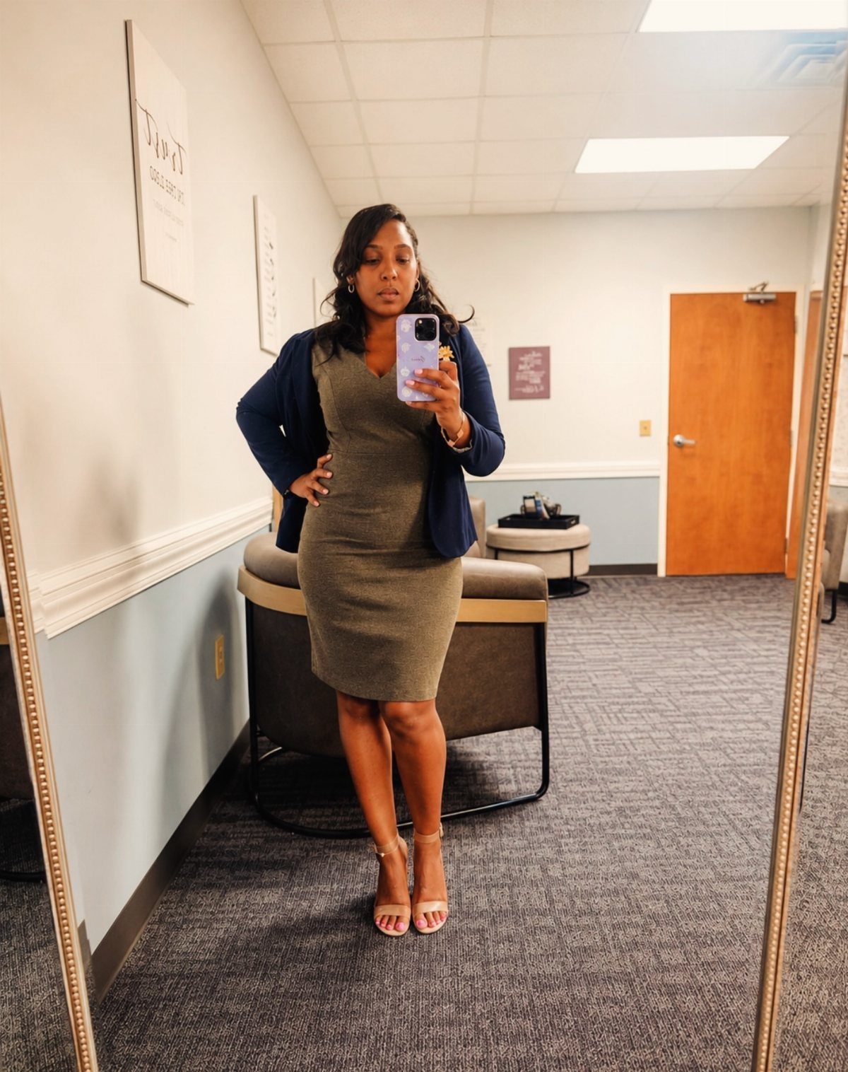 Woman in olive green sheath dress and navy blazer posing in office mirror