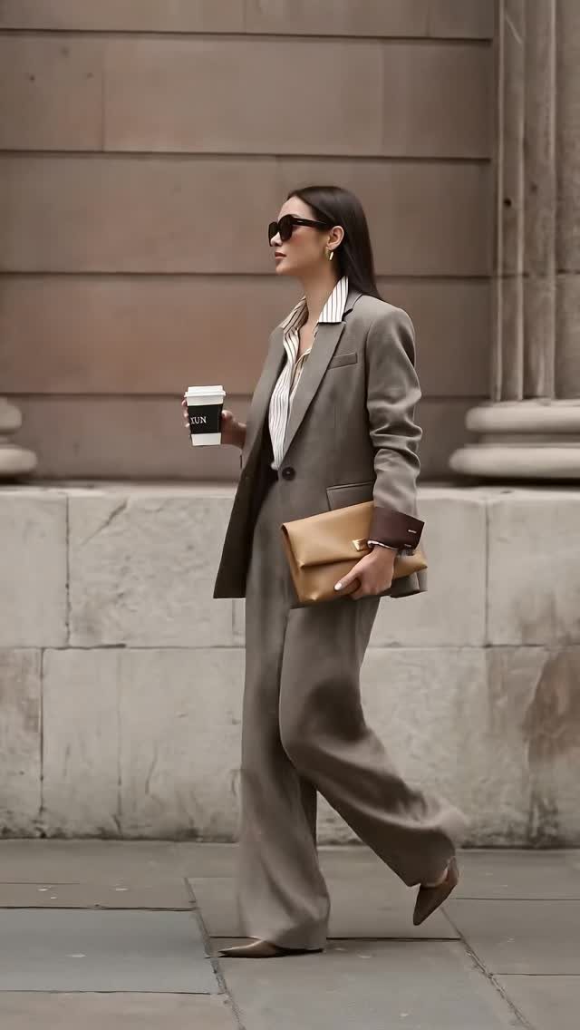 Woman in a taupe tailored suit with wide-leg trousers and a tan clutch bag.