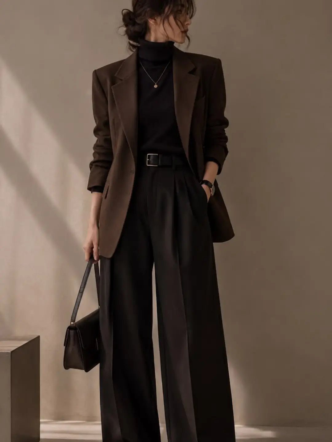 Woman in brown blazer, black turtleneck, and wide-leg trousers holding a black handbag.