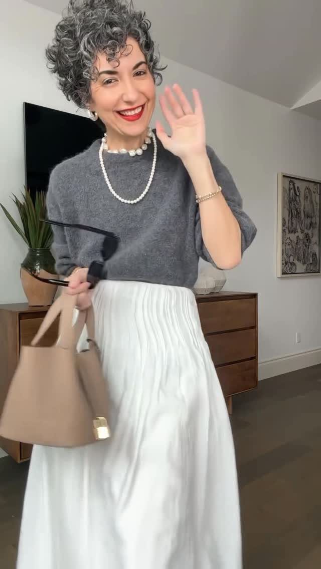8 Fall Outfits for Women Over 50 to Wear Right Now 3 Woman with grey hair wearing a grey sweater and white pleated midi skirt.