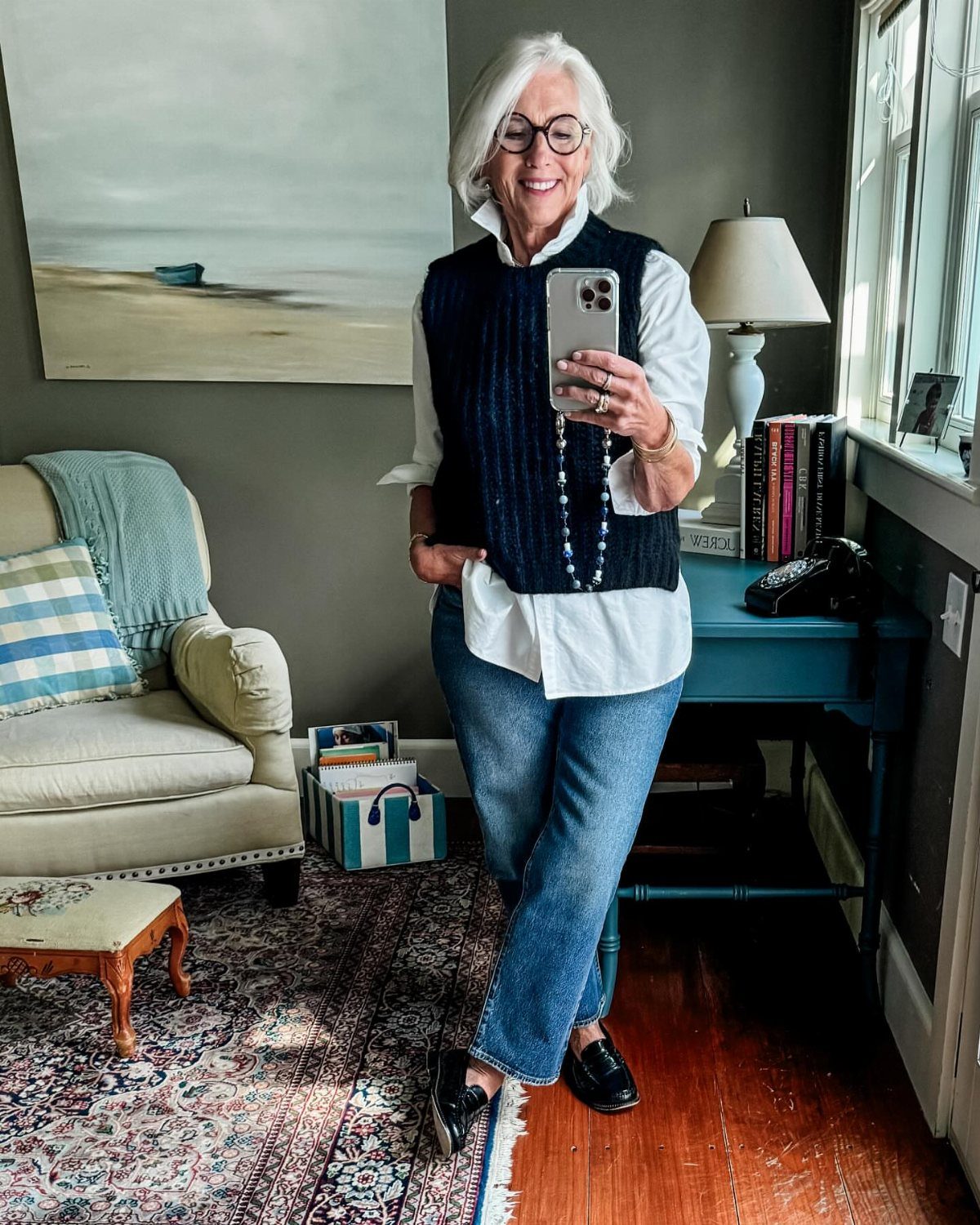 8 Fall Outfits for Women Over 50 to Wear Right Now 2 Woman with white hair wearing a navy sweater vest over a white shirt and jeans.