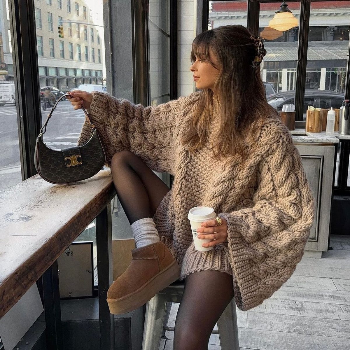 Fall Outfits for Women: 9 Ideas From Denim to Burgundy 7 Woman in oversized brown cable-knit sweater and tan suede platform boots sitting by window.
