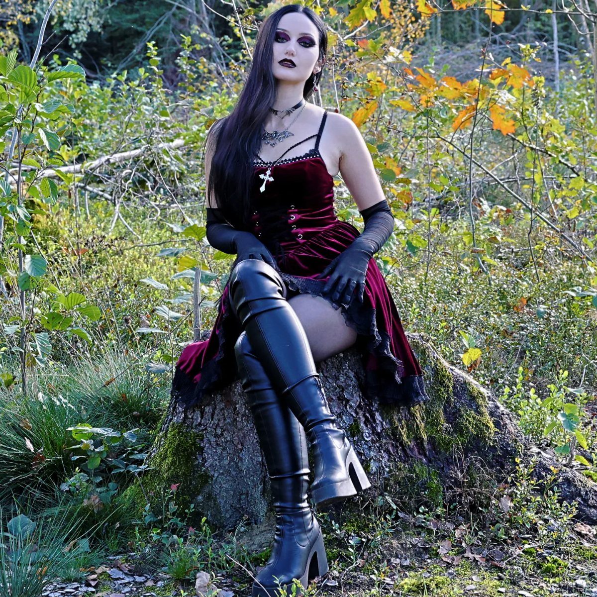 Woman in burgundy velvet dress and black leather boots sitting on a tree stump.