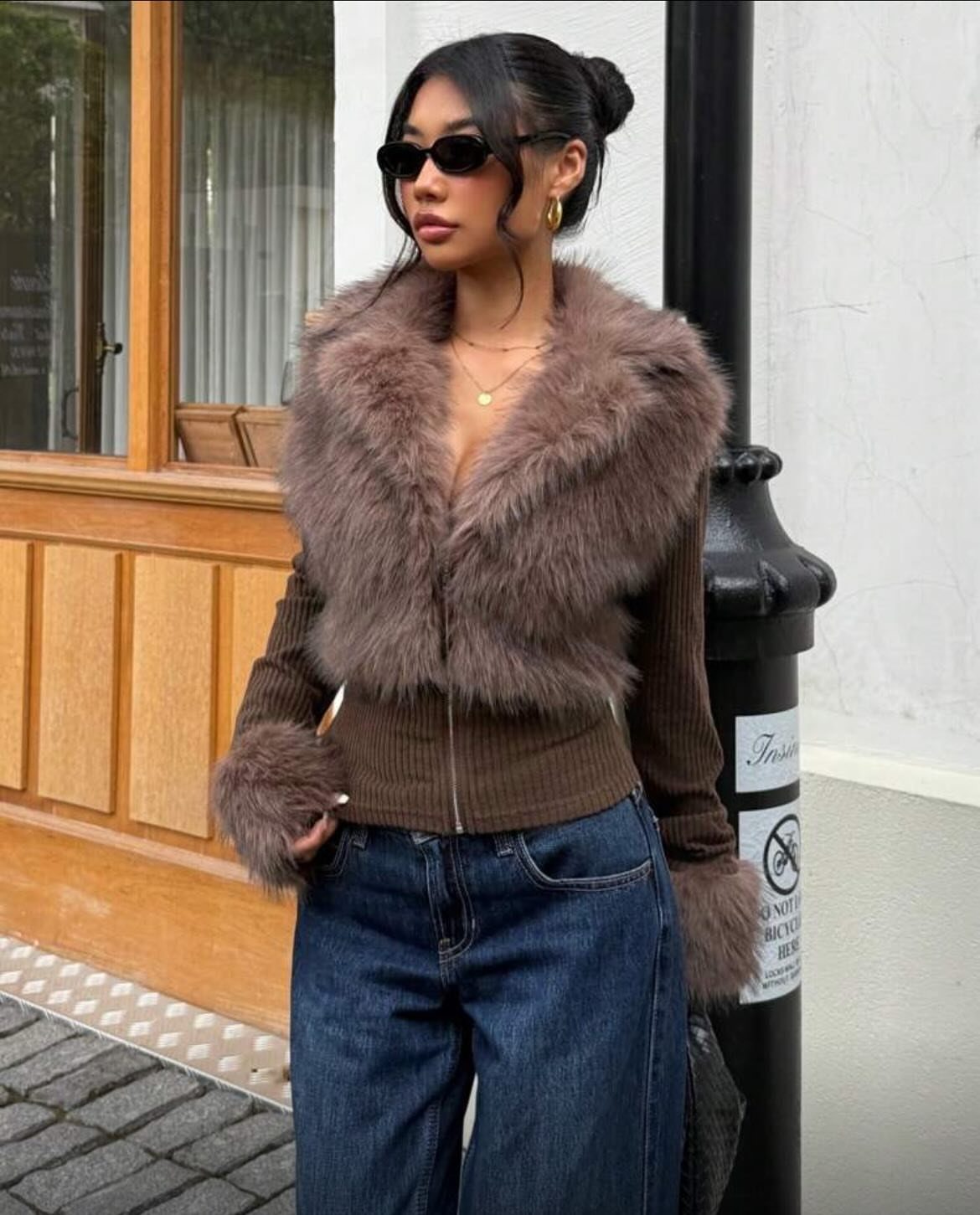 Woman in a brown faux-fur collar cropped jacket and blue jeans posing outdoors.