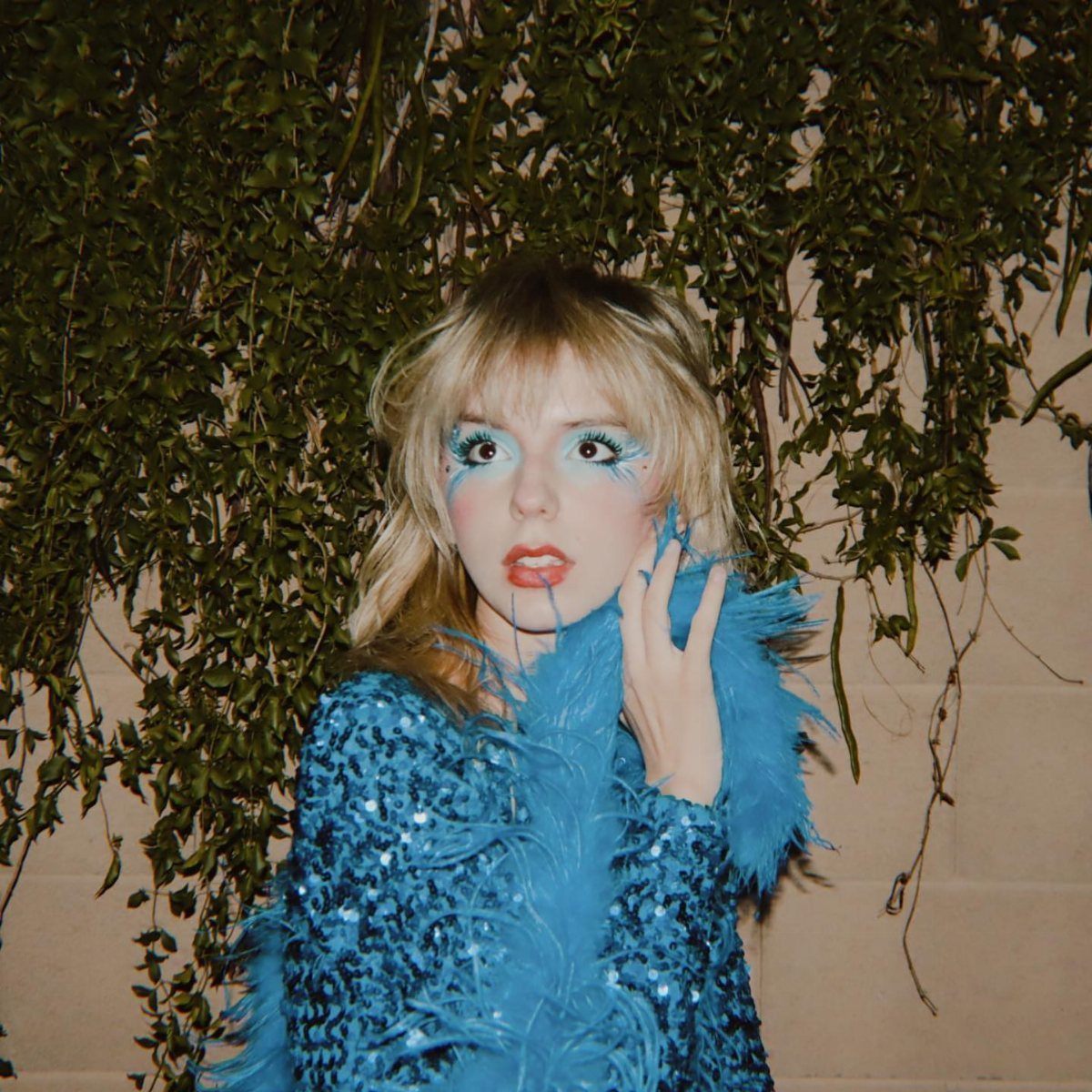 Person in blue sequin clothing and a blue feather boa against green ivy.