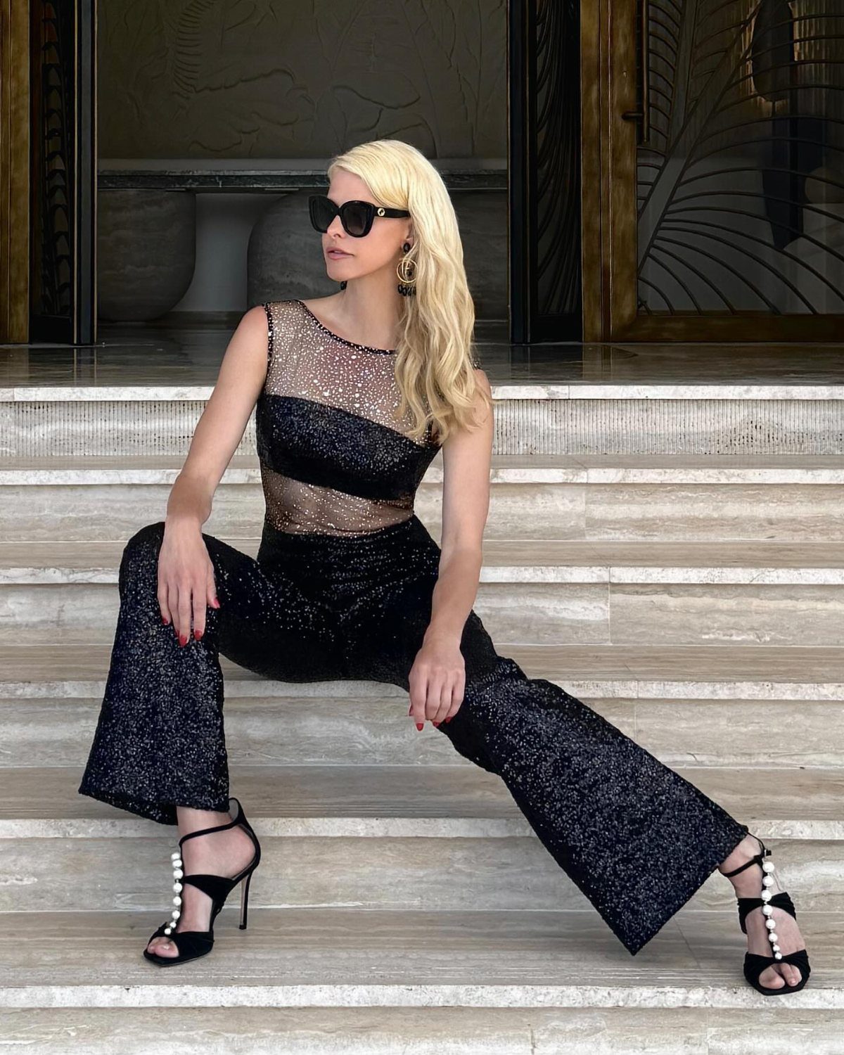 Woman in a black sequined jumpsuit with sheer bodice sitting on white marble stairs