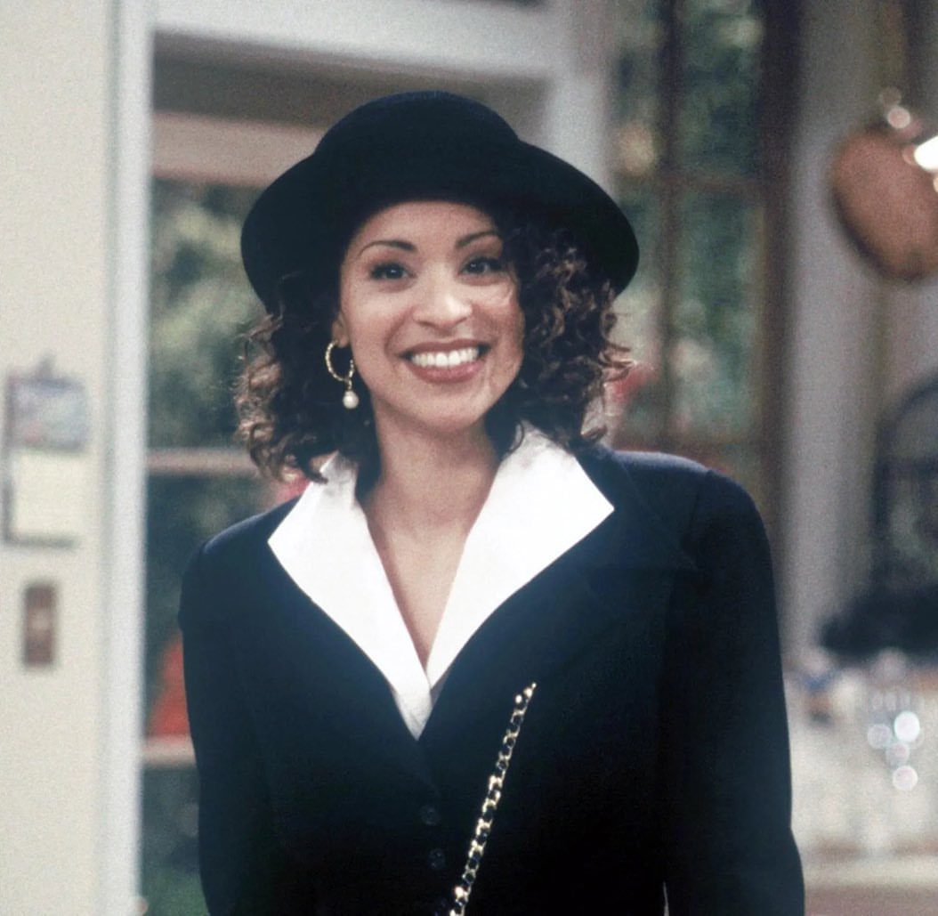 Hilary Banks Outfits: 12 Iconic Looks to Recreate Her Style 10 Woman wearing a black hat, black blazer, and white shirt smiling indoors.