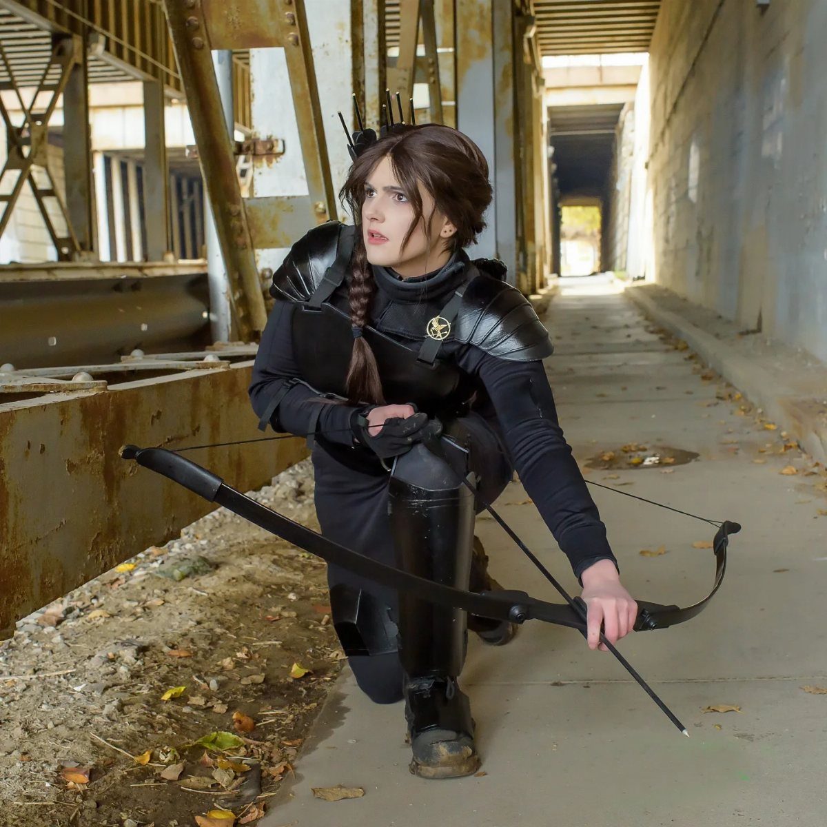 Hunger Games Outfits: 8 Iconic Looks from the Films 1 Person in black dystopian huntress costume with bow kneeling in industrial setting.
