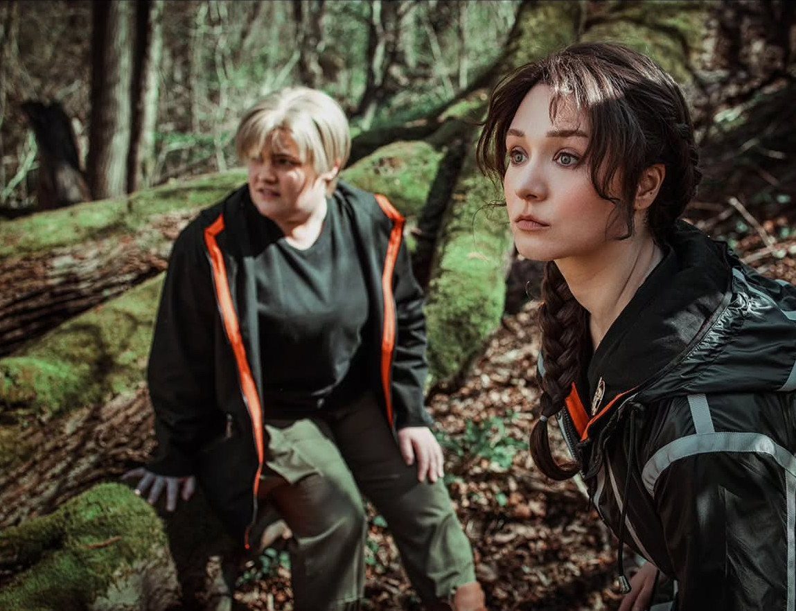 Hunger Games Outfits: 8 Iconic Looks from the Films 7 Two women wearing black and orange tactical outfits in a mossy forest setting.