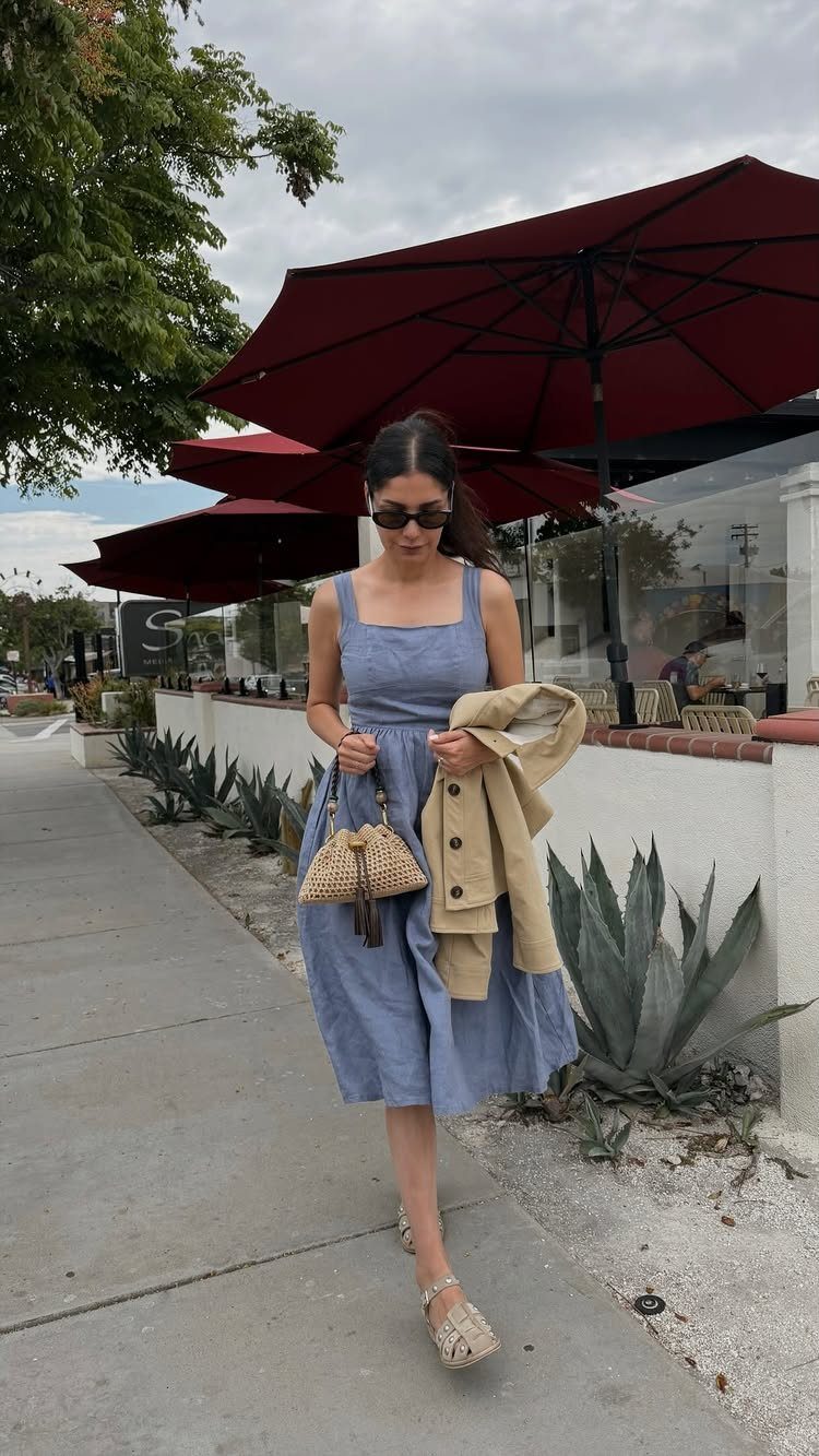 Woman in a blue linen midi dress walking past red cafe umbrellas.