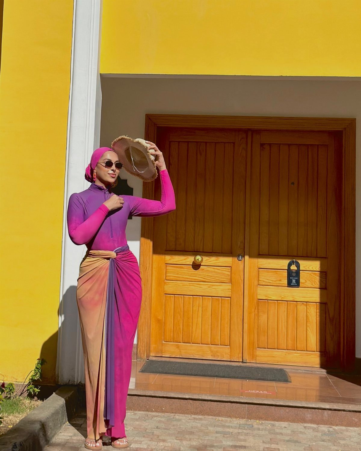 Woman in purple long-sleeved top and wrap skirt holding a straw hat.