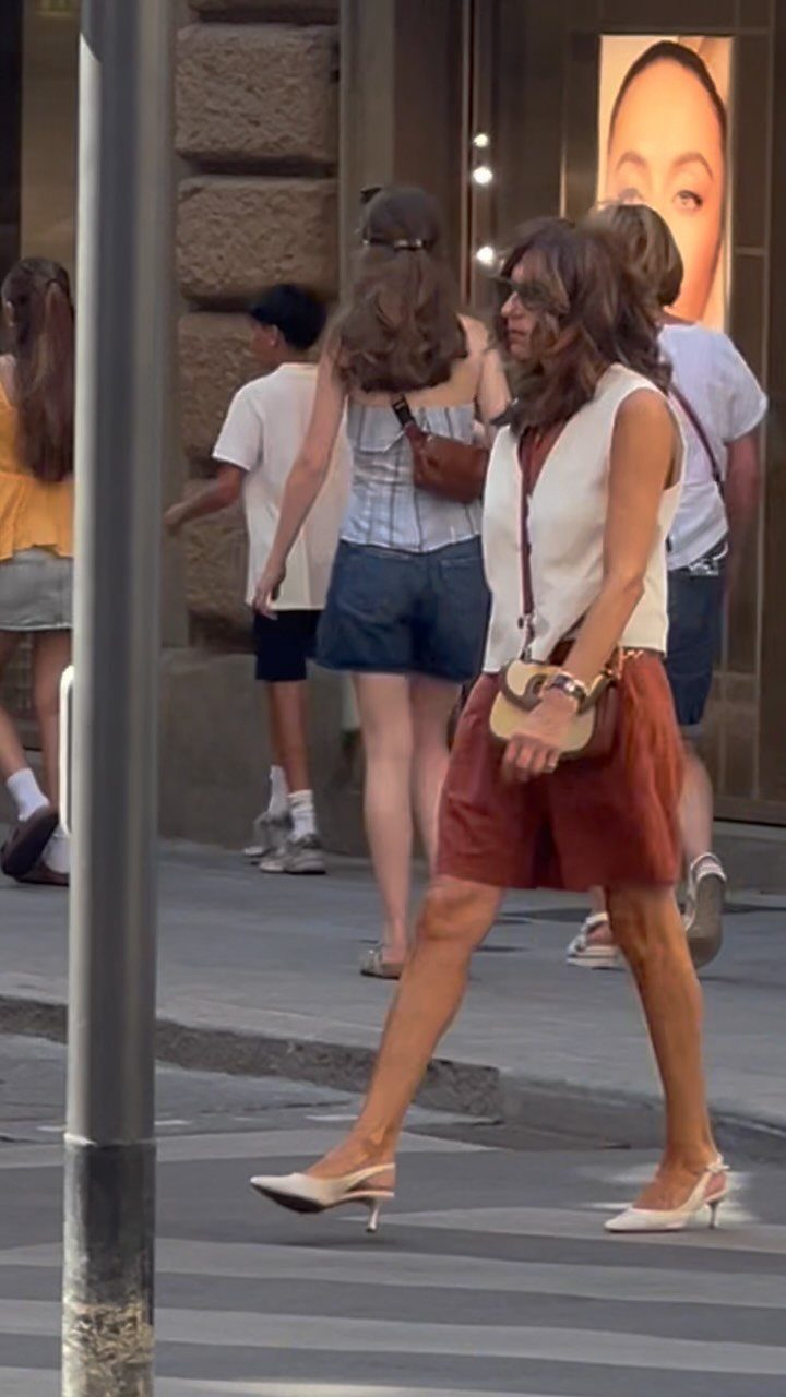 Woman wearing reddish-brown tailored shorts, white top, and white heels walking on street.