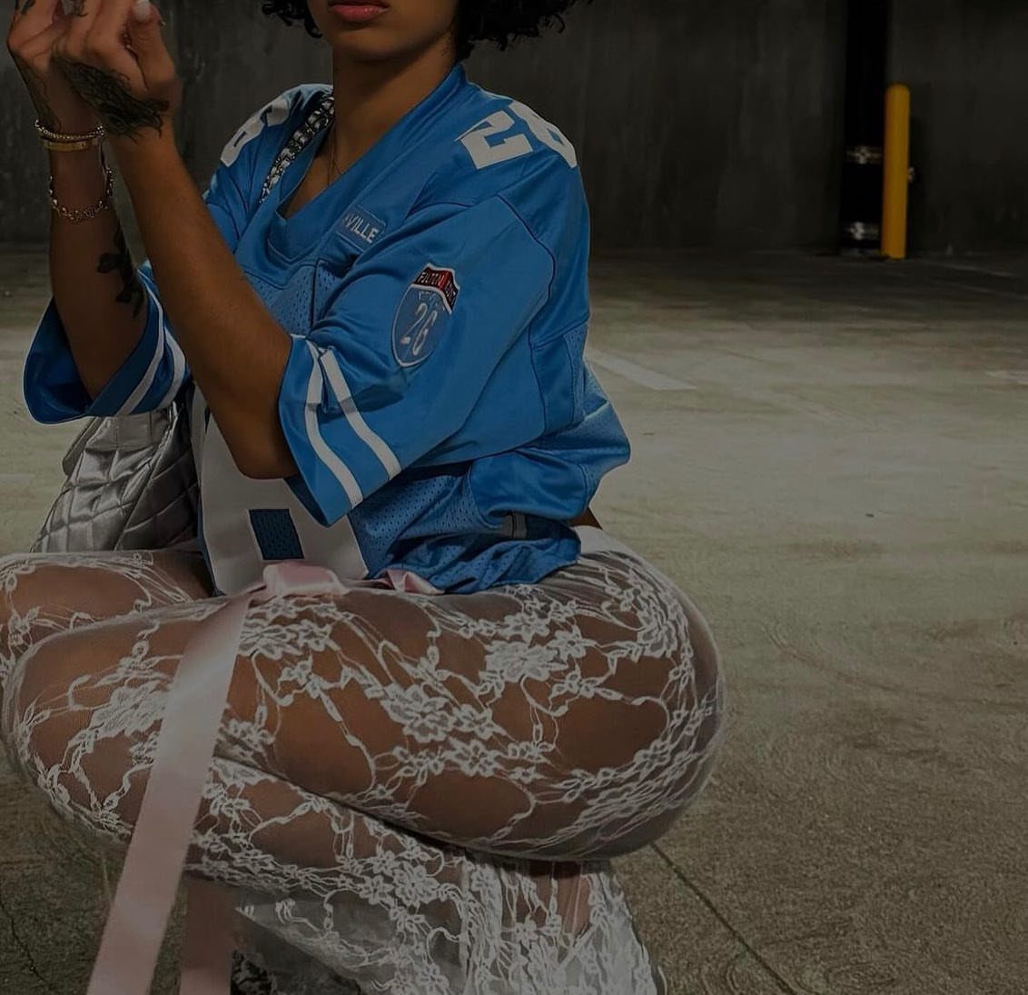 Kali Uchis Outfits: 12 Bold Looks for Retro Style 11 Person in blue sports jersey and white lace leggings crouching in a garage.