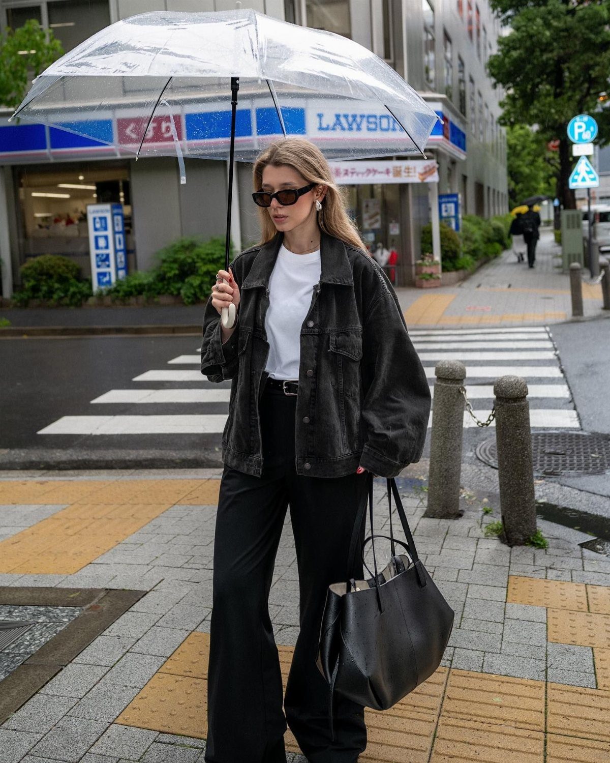 Woman in oversized black denim jacket and black trousers holding a clear umbrella.