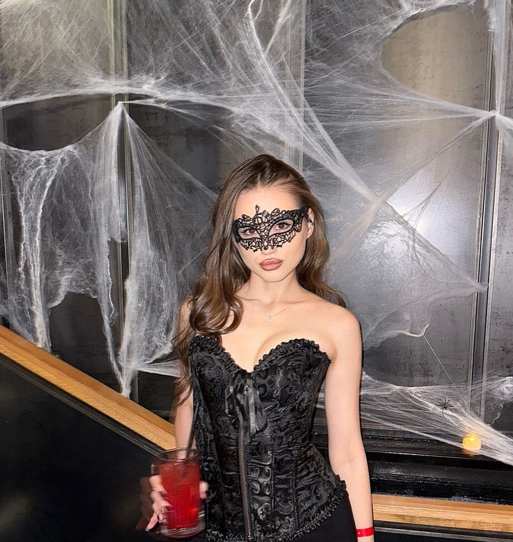 Woman in black lace corset and masquerade mask posing before giant spider webs.