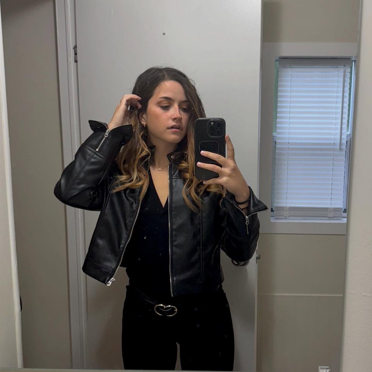 Woman wearing a black leather jacket and black trousers taking a mirror selfie.