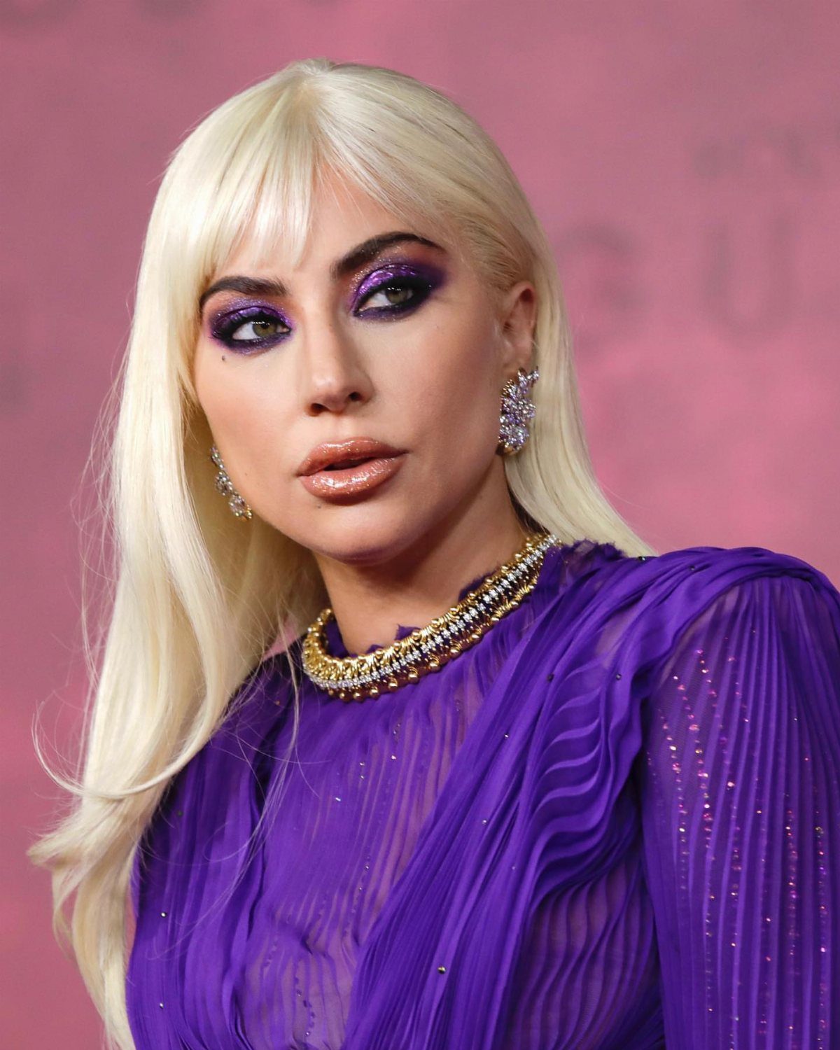 11 Lady Gaga Outfits That Defined Her Style Evolution 4 Lady Gaga wearing a pleated purple dress and gold necklace against a pink background