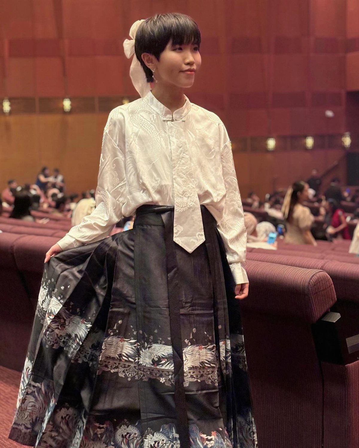 13 Laufey Concert Outfit Ideas for a Whimsical Night Out 5 Person wearing a white high-collar blouse and a black patterned maxi skirt in a theater.