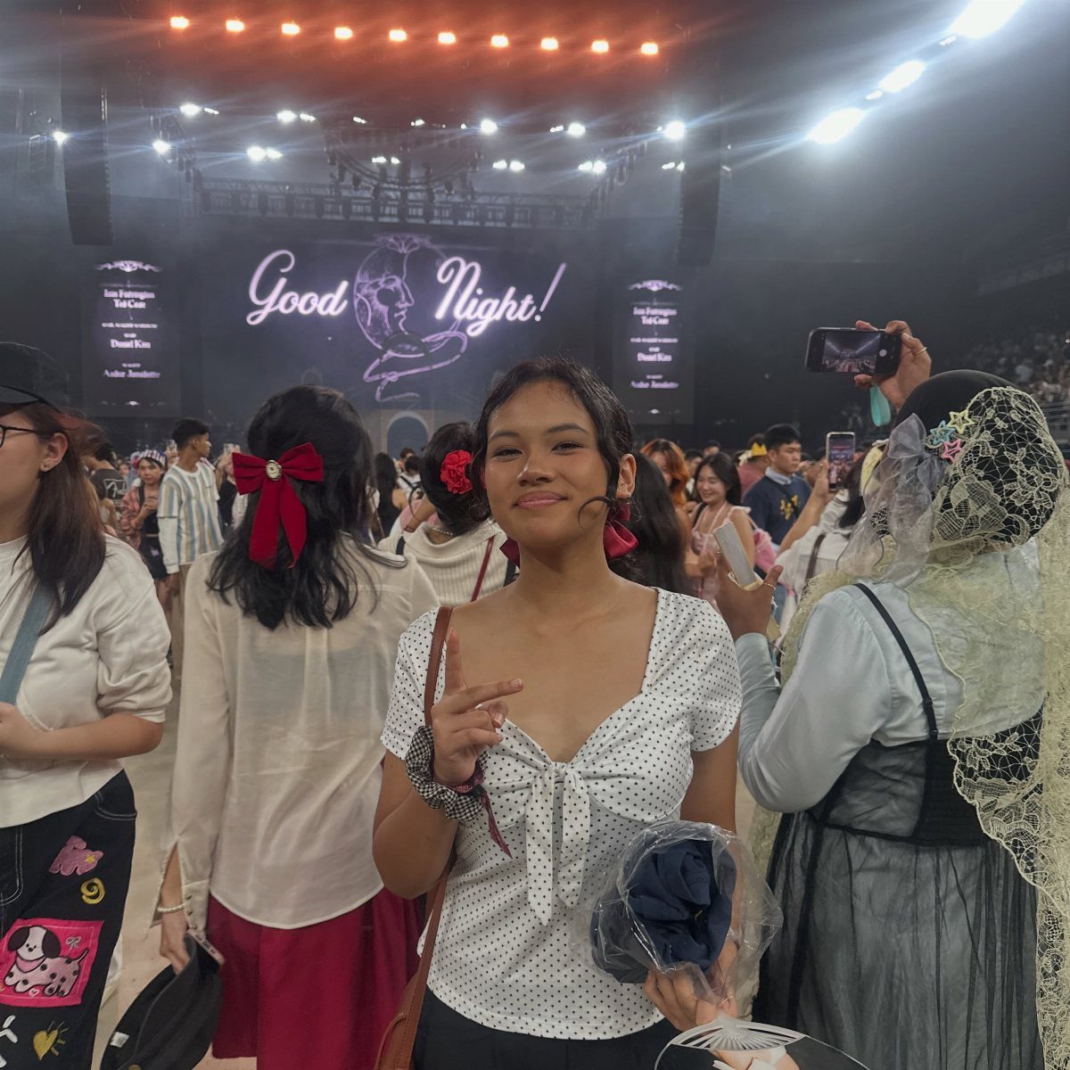 13 Laufey Concert Outfit Ideas for a Whimsical Night Out 13 Young woman in a white polka-dot blouse posing at a Laufey concert venue.
