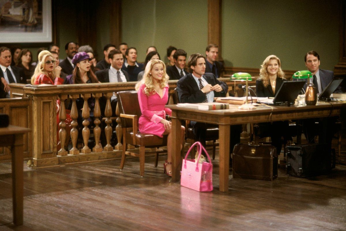 10 Legally Blonde Outfits to Channel Elle Woods in 2026 4 Blonde woman wearing a bright pink dress sitting in a courtroom setting.