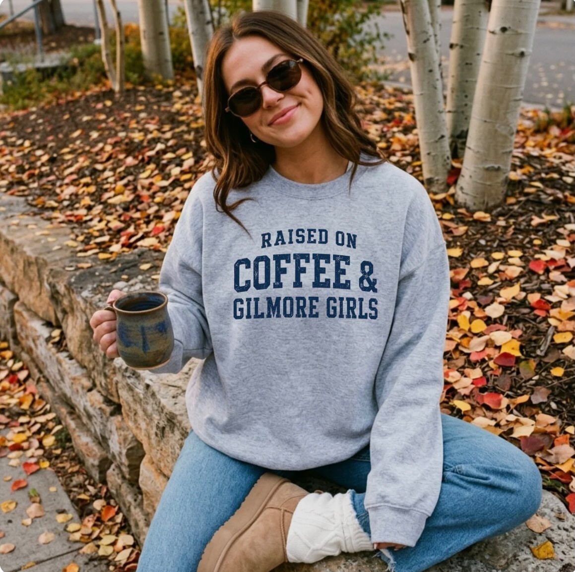 11 Lorelai Gilmore Outfits to Recreate Her Iconic Style 3 Woman in a grey Gilmore Girls graphic sweatshirt sitting on a stone wall outdoors.