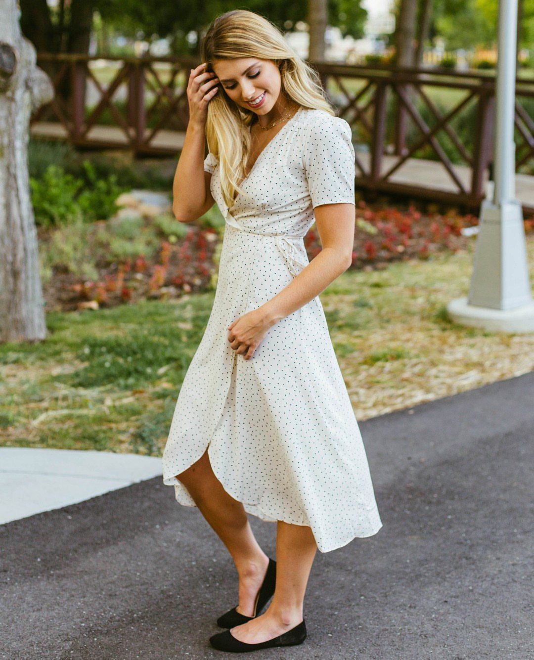11 Lorelai Gilmore Outfits to Recreate Her Iconic Style 8 Blonde woman wearing a cream polka dot wrap dress and black flat shoes outdoors.