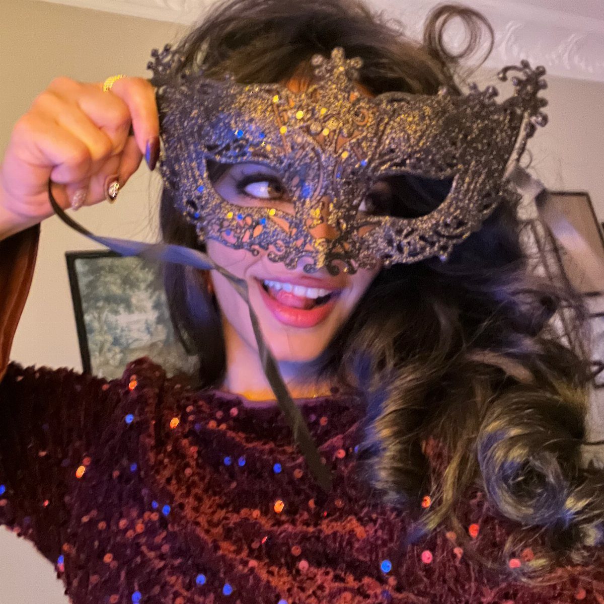 10 Stunning Masquerade Outfits for Women to Wear This Year 5 Woman wearing a burgundy sequined top and holding a gold filigree masquerade mask.
