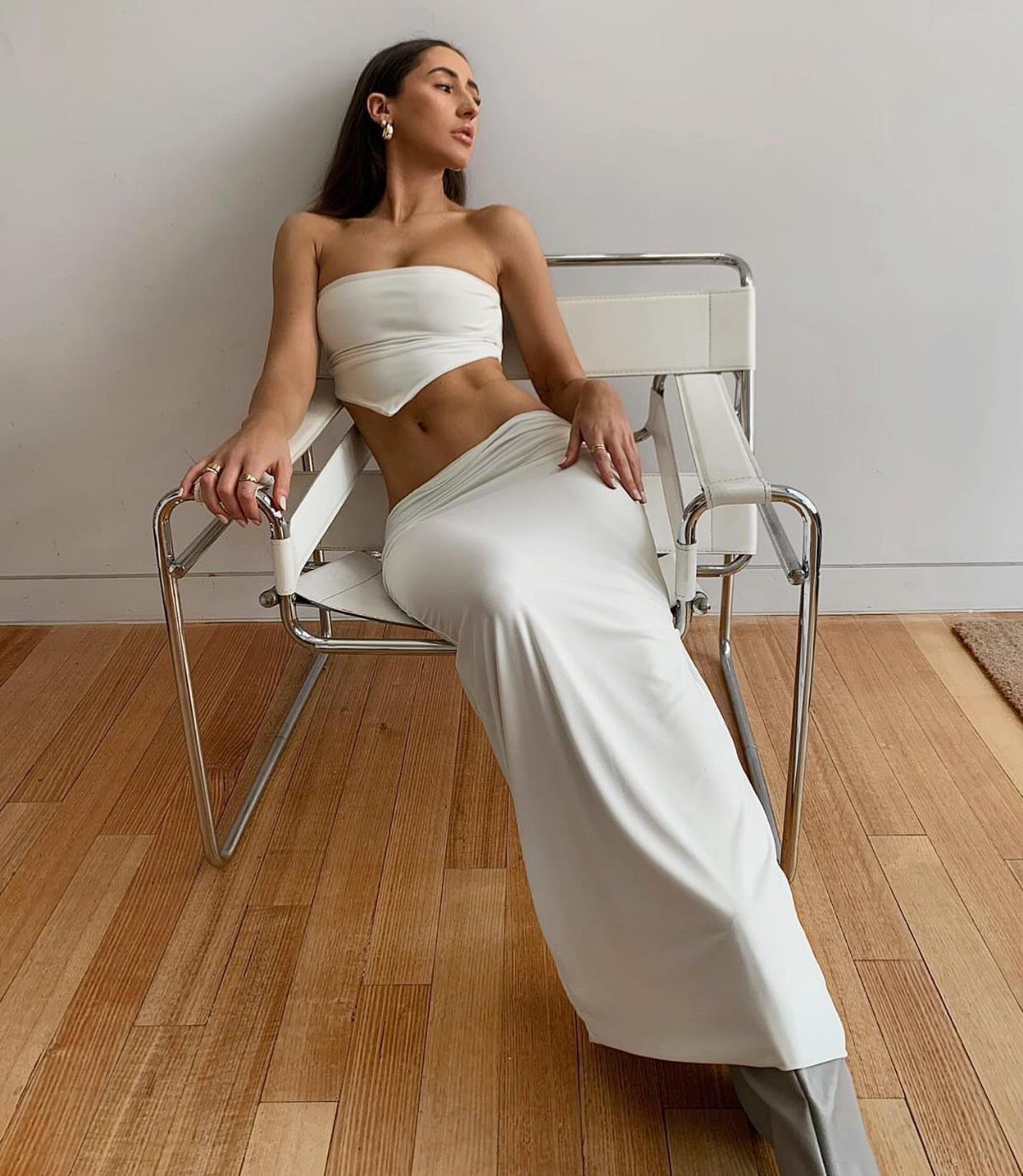 12 Maxi Skirt Outfits to Wear All Year in 2026 9 Woman in white strapless top and low-rise white maxi skirt sitting in chrome chair.