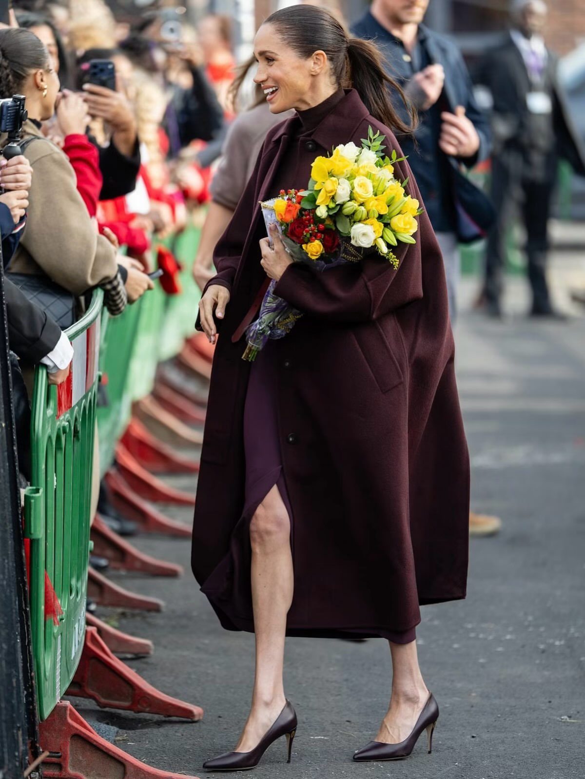 Meghan Markle Outfits: 17 Ideas from Tailored to Casual 1 Meghan Markle in a burgundy tailored coat and dress holding yellow flowers.