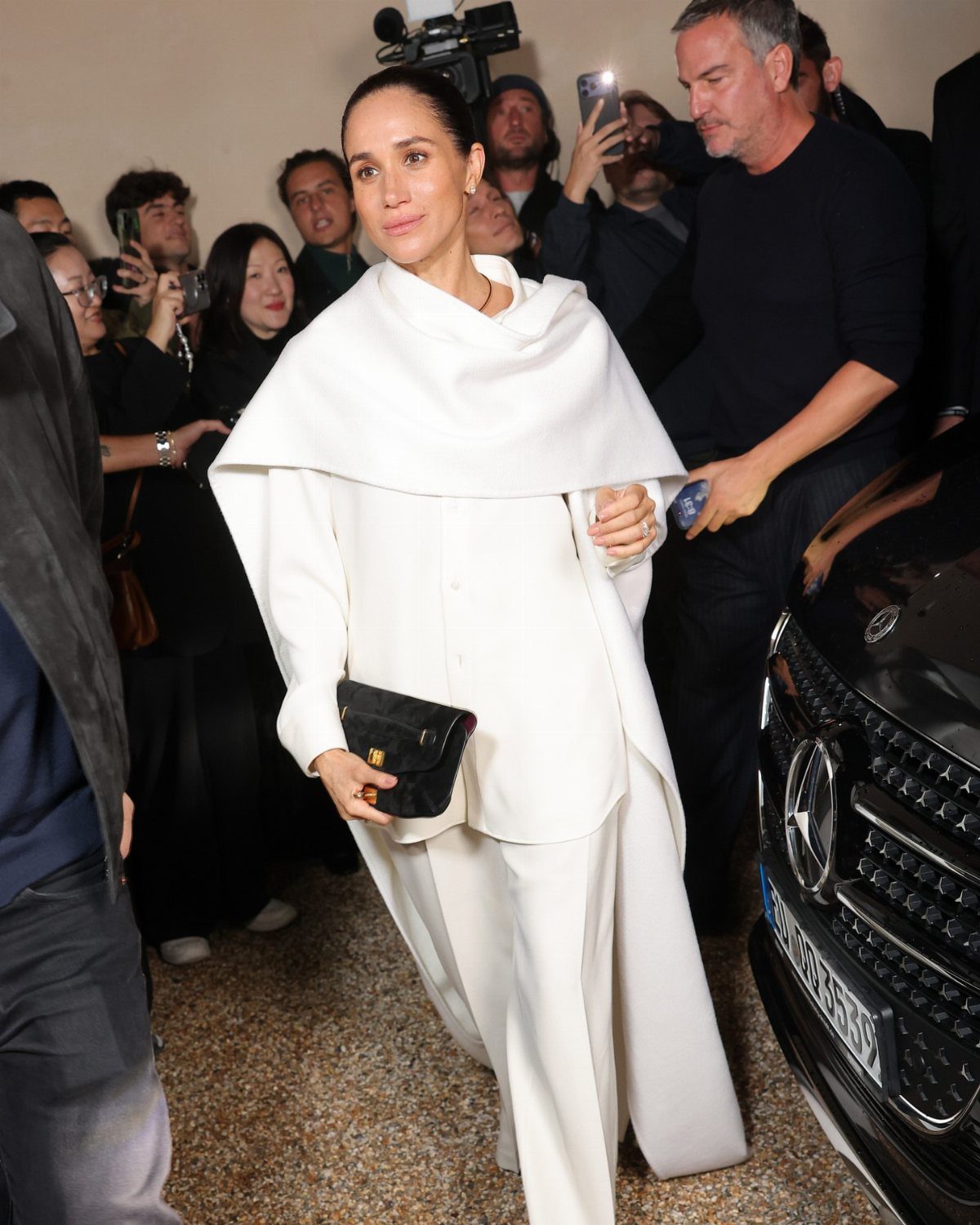 Meghan Markle Outfits: 17 Ideas from Tailored to Casual 7 Meghan Markle wearing a structured white cape and trousers with a black clutch.