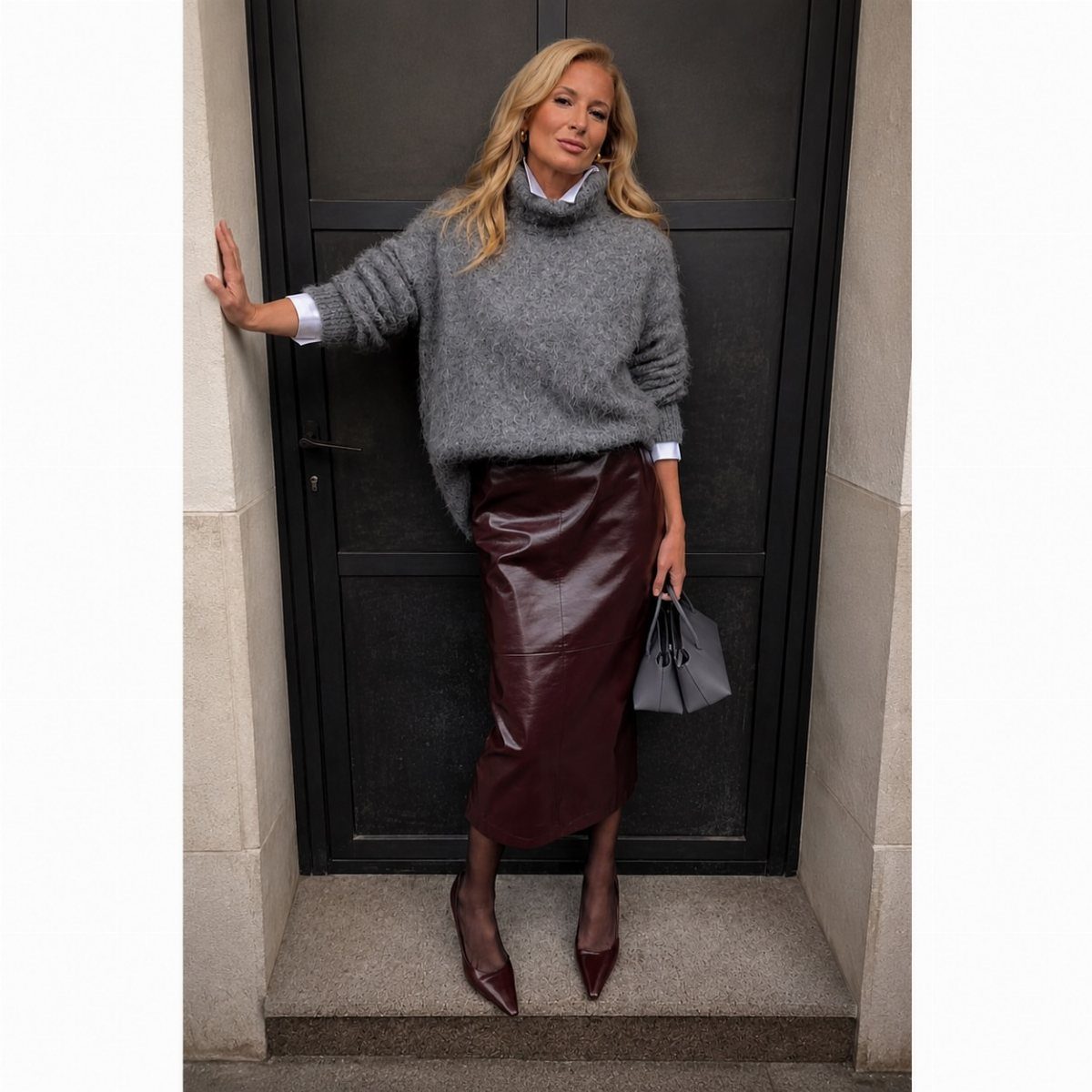12 Midi Skirt Outfits for Every Season and Style 7 Woman in grey oversized sweater and burgundy leather midi skirt posing by a door.