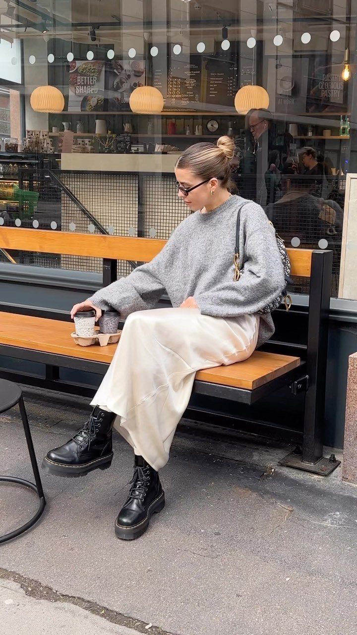 12 Midi Skirt Outfits for Every Season and Style 3 Woman in grey knit jumper, cream satin slip skirt, and black biker boots sitting outdoors.