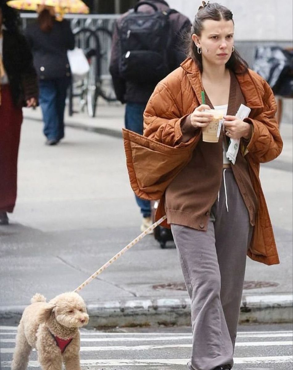 15 Millie Bobby Brown Outfits to Inspire Your Wardrobe 7 Millie Bobby Brown walking a poodle in a brown oversized jacket and grey trousers.