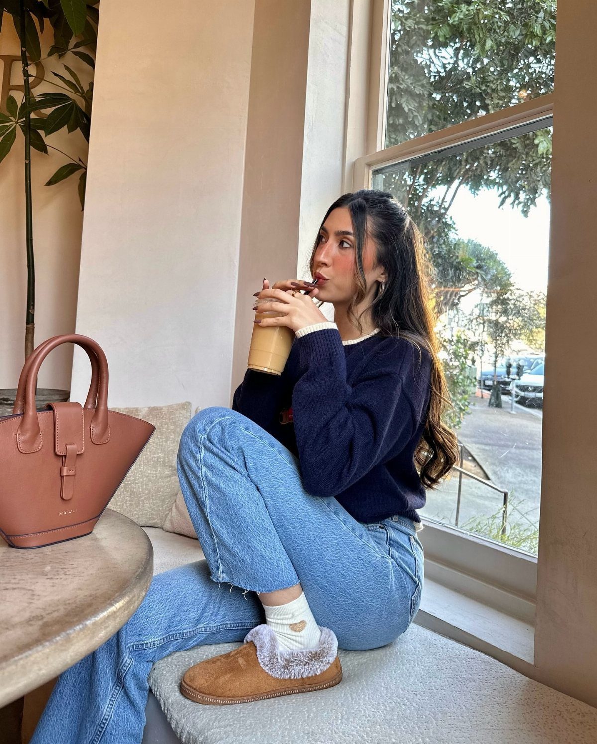 15 Millie Bobby Brown Outfits to Inspire Your Wardrobe 4 Woman in navy sweater and blue jeans sitting by a window with a handbag.