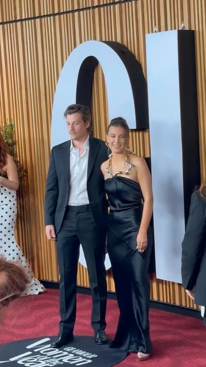 15 Millie Bobby Brown Outfits to Inspire Your Wardrobe 5 Man in black suit and woman in black satin gown posing on red carpet.