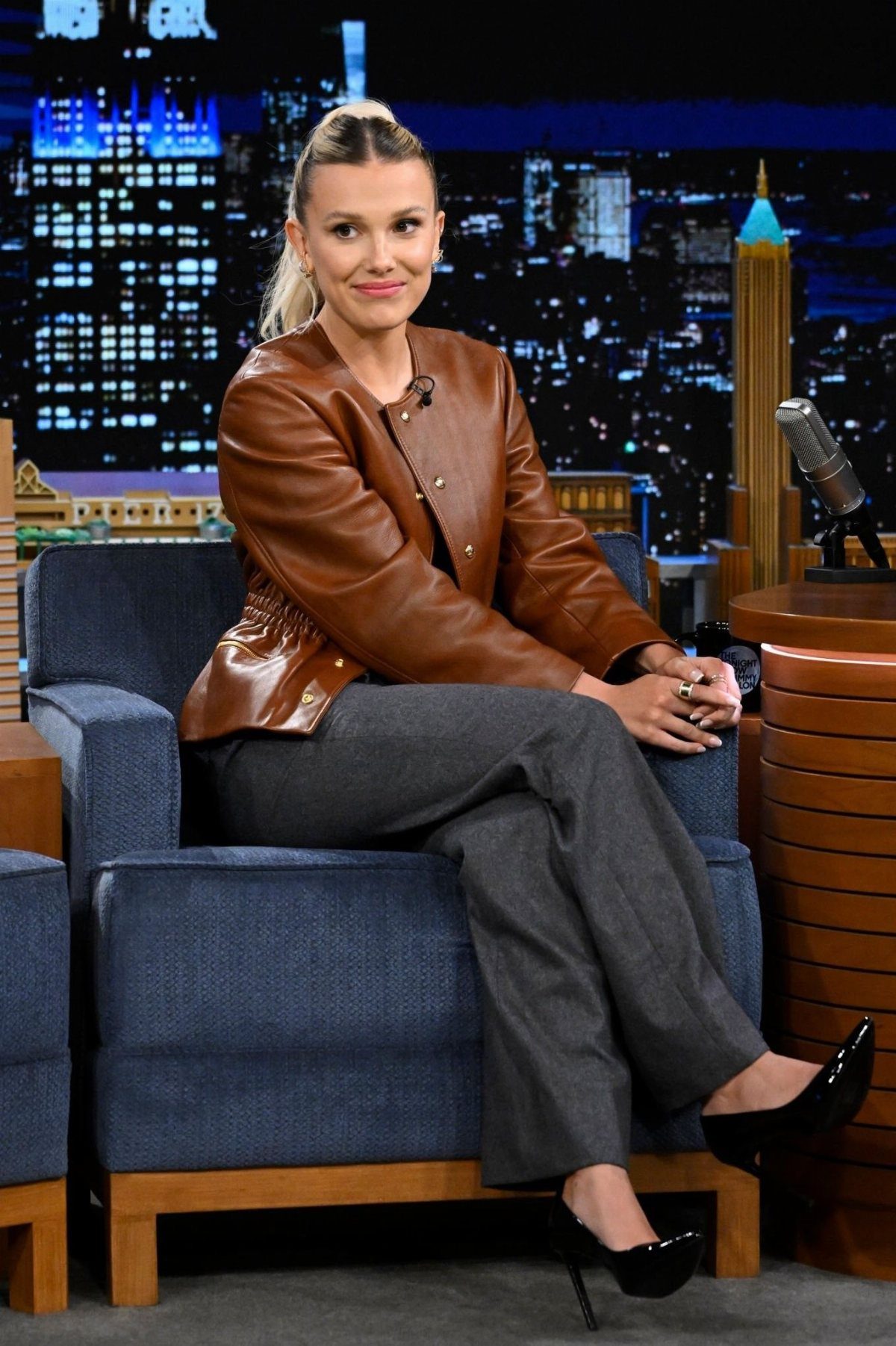 15 Millie Bobby Brown Outfits to Inspire Your Wardrobe 3 Millie Bobby Brown wearing a brown leather jacket and grey tailored trousers sitting down.