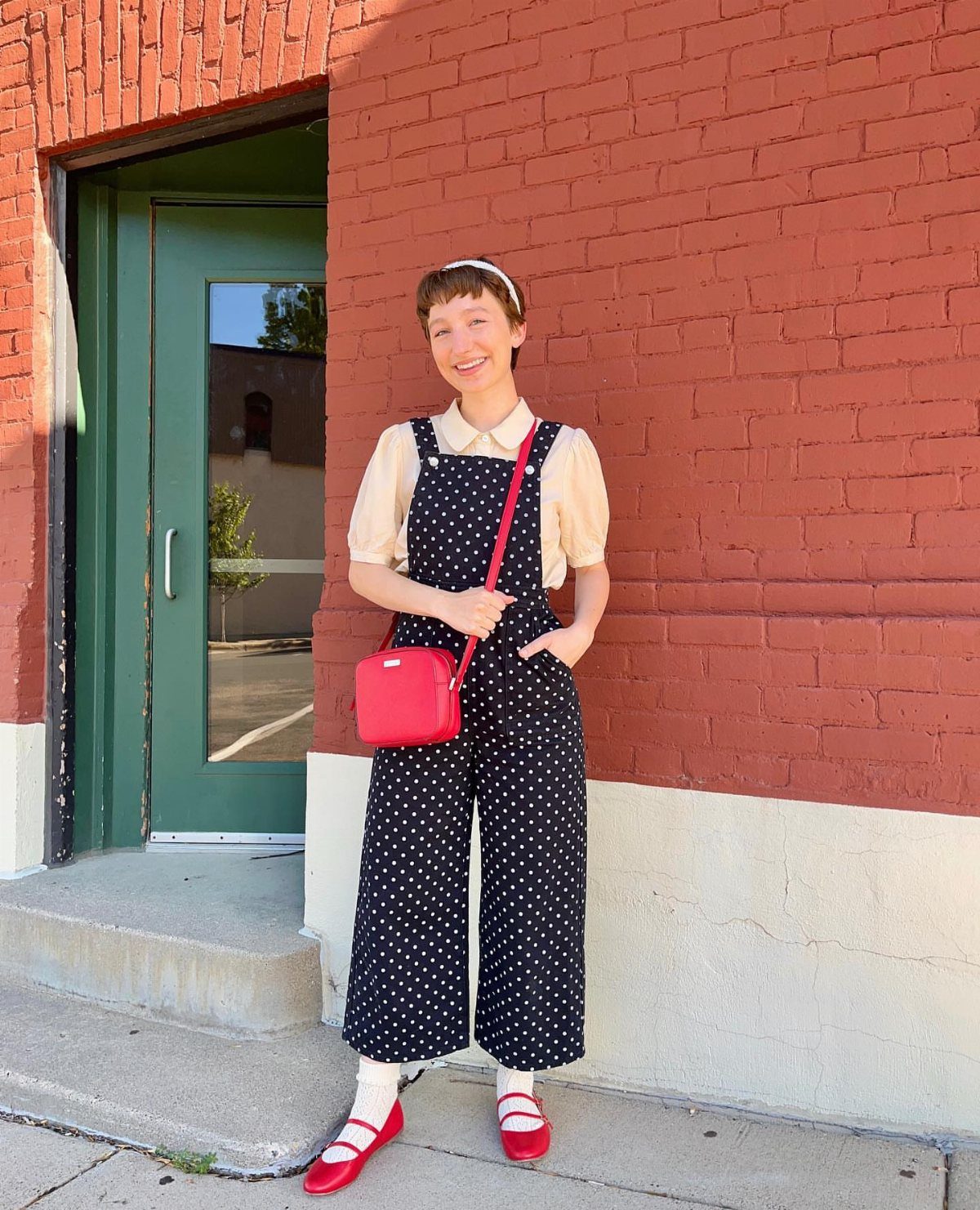 15 Millie Bobby Brown Outfits to Inspire Your Wardrobe 2 Woman in polka dot jumpsuit and red accessories posing by a red brick wall.