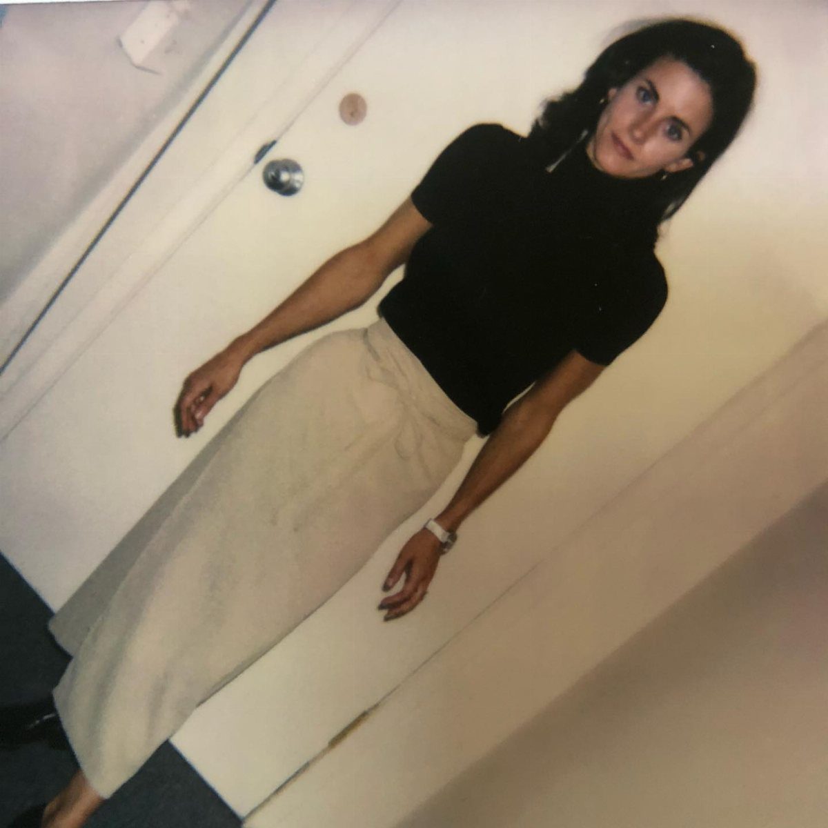 Monica Geller Outfits: 8 Iconic Looks to Recreate Now 7 Woman wearing a black short-sleeved turtleneck and a long beige skirt against a wall.