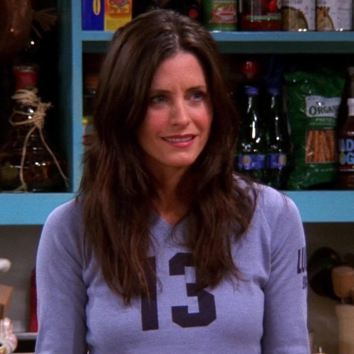 Monica Geller Outfits: 8 Iconic Looks to Recreate Now 6 Monica Geller wearing a purple long-sleeved t-shirt with the number 13 in a kitchen.