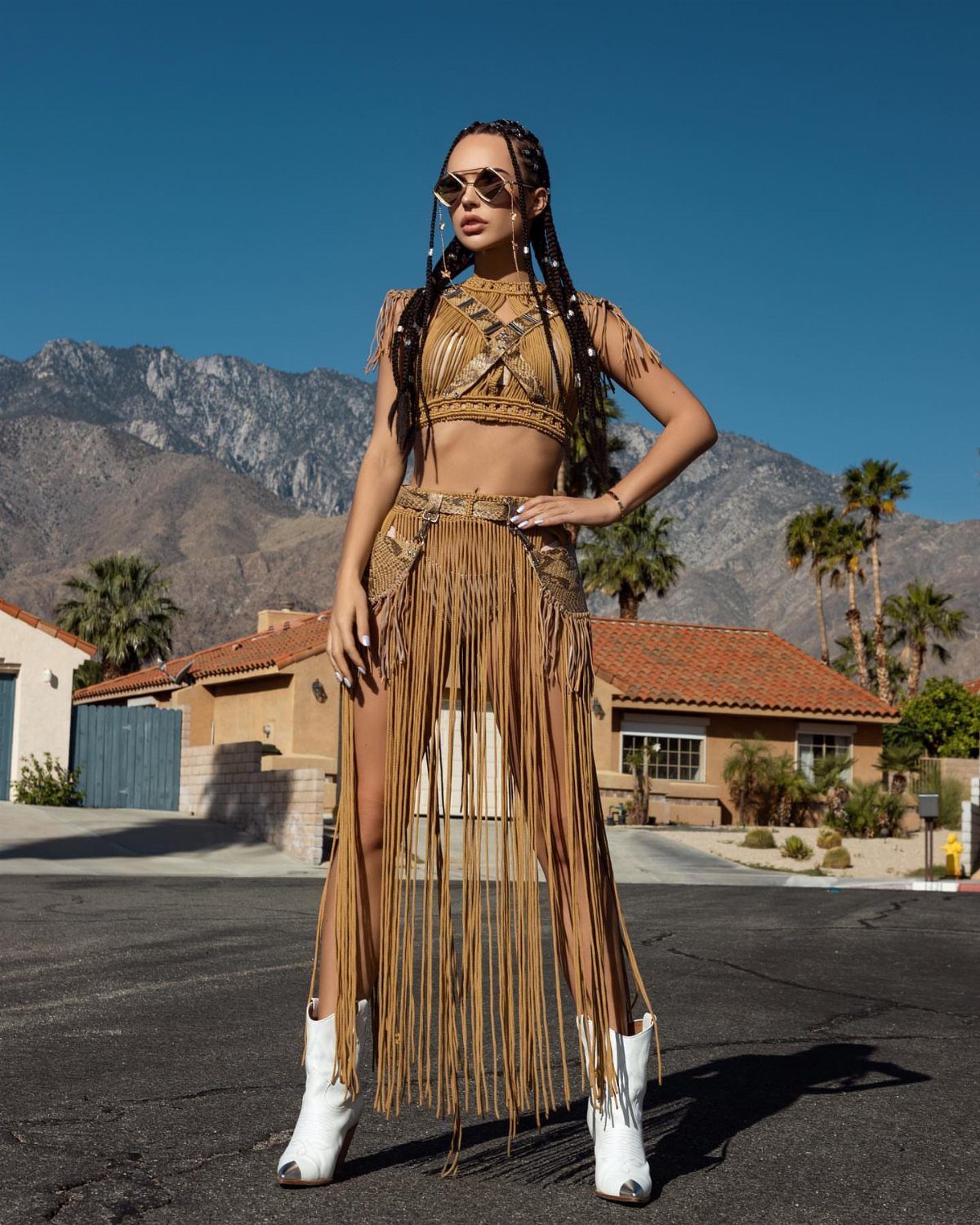9 Music Festival Outfits for Every Weather and Vibe in 2026 5 Woman in tan fringe outfit and white cowboy boots posing against mountain backdrop.