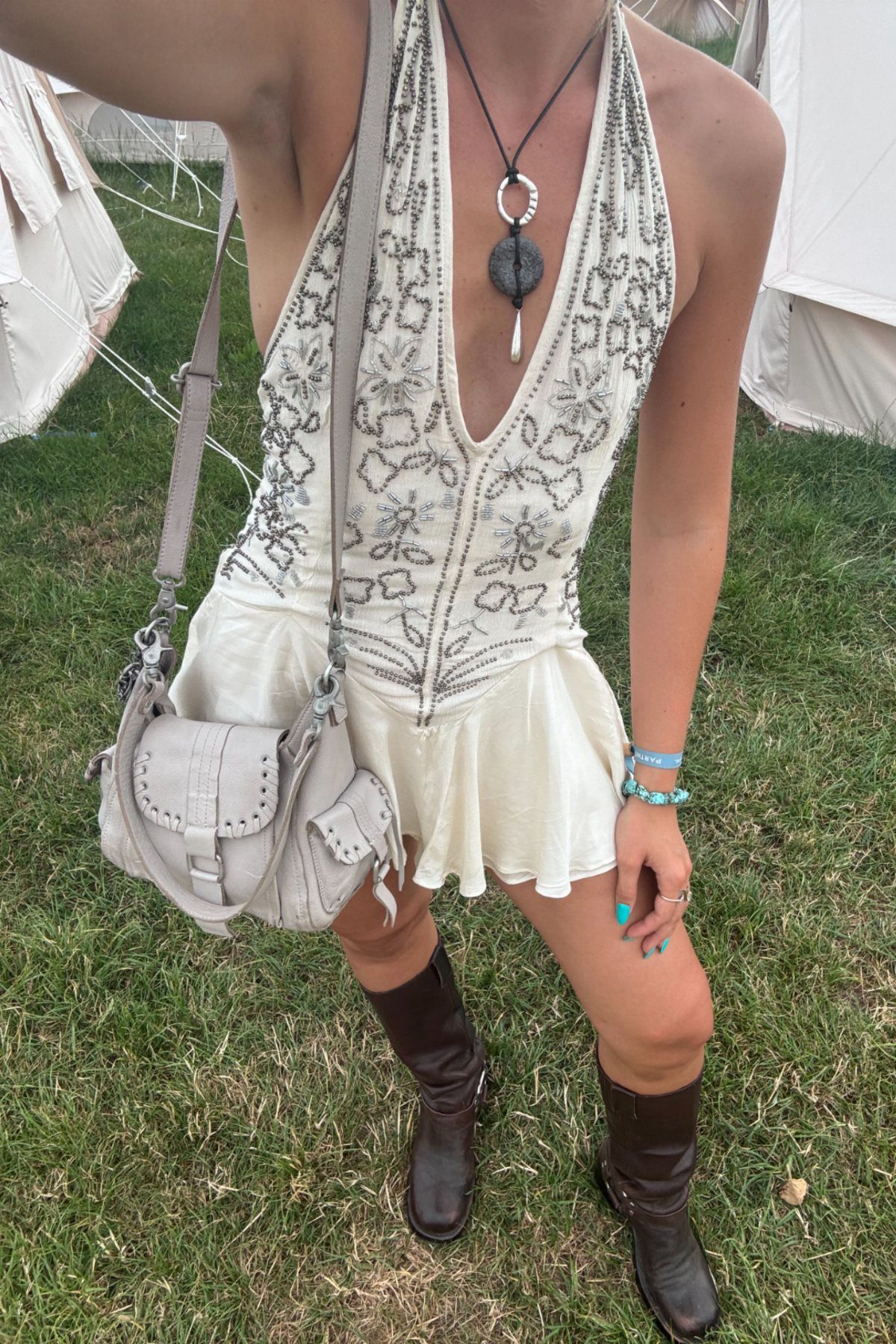 9 Music Festival Outfits for Every Weather and Vibe in 2026 6 Woman wearing a white halterneck dress, brown biker boots, and a grey woven bag.