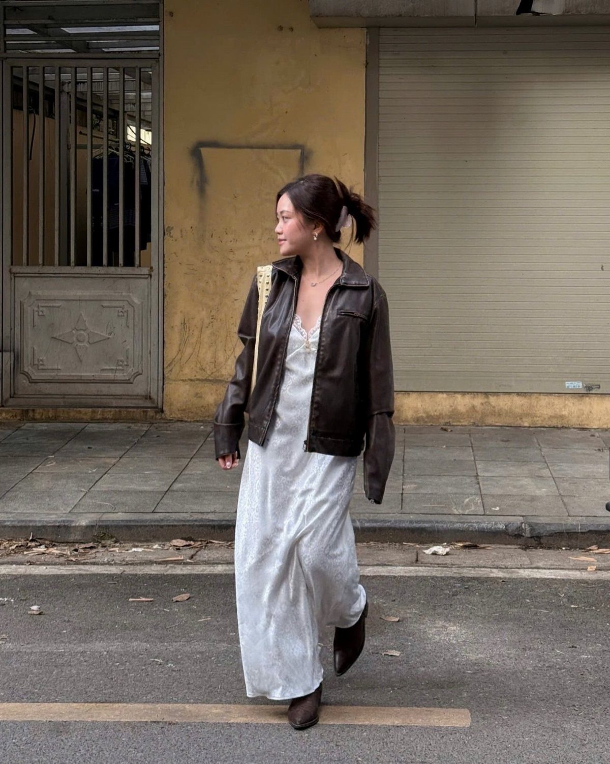 9 Music Festival Outfits for Every Weather and Vibe in 2026 4 Woman wearing a white maxi dress, brown leather jacket, and brown boots on street.