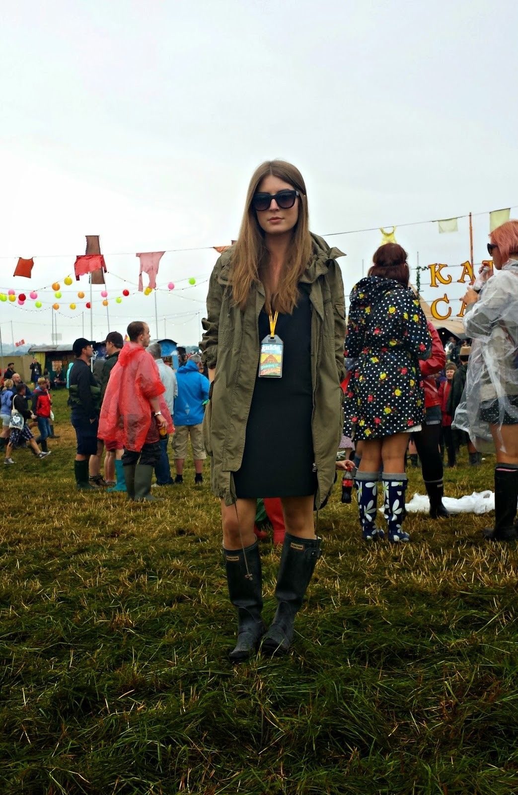 9 Music Festival Outfits for Every Weather and Vibe in 2026 1 Woman wearing olive rain jacket, black bodycon dress, and gumboots at a festival.
