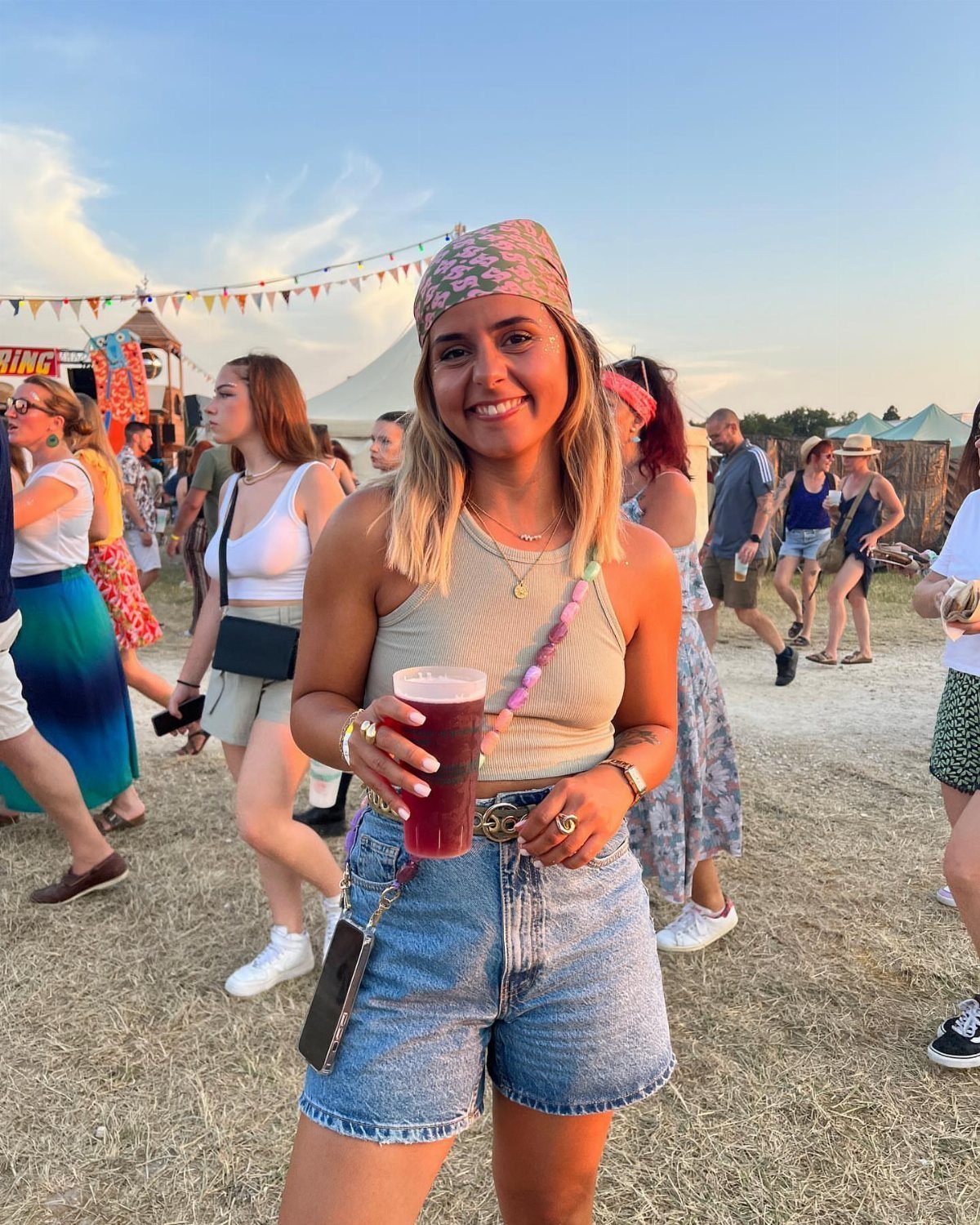 9 Music Festival Outfits for Every Weather and Vibe in 2026 7 Woman in beige tank top and denim shorts smiling at an outdoor music festival.