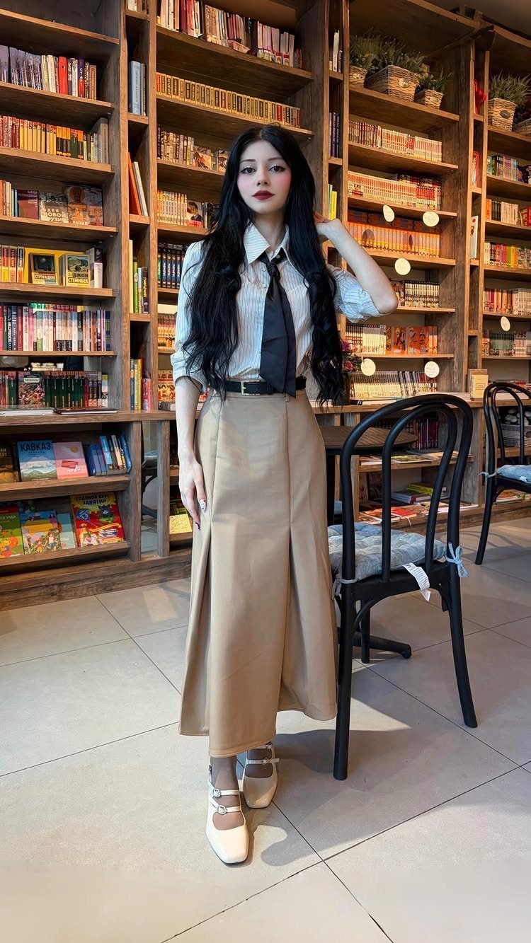 8 Office Siren Outfits to Master the Look in 2026 7 Woman in a white shirt, black tie, and tan skirt posing in a library.