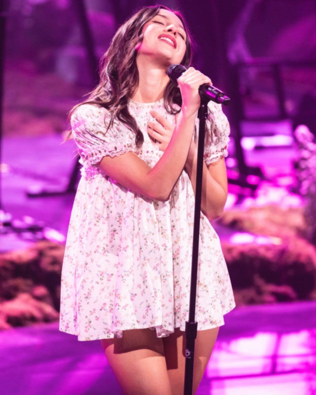 Woman singing on stage wearing a white floral babydoll dress under purple lights