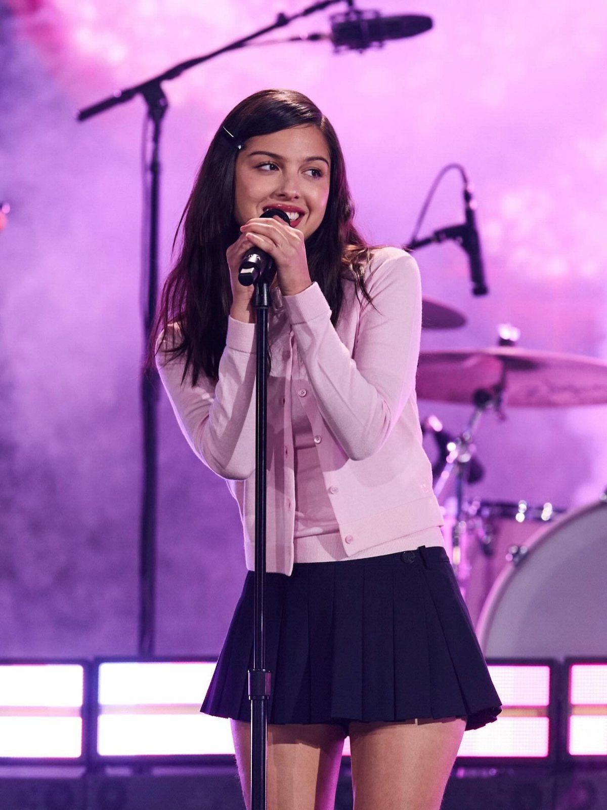 Olivia Rodrigo singing on stage wearing a pink cardigan and black pleated skirt.