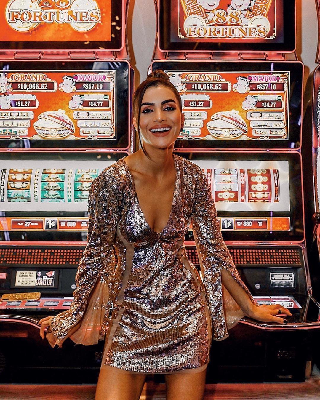 Woman in a gold sequined cocktail dress posing before casino slot machines.