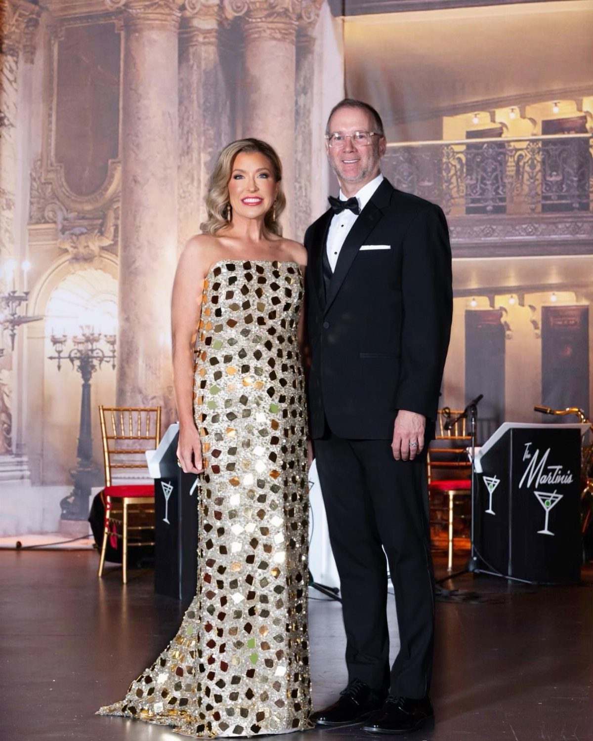 Woman in gold sequined floor-length gown and man in black tuxedo posing together.